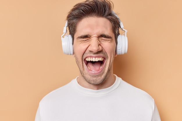 Emotional brunet man exclaims loudly keeps mouth opened dressed in casual white jumper listens music via wireless headphones has eyes shut isolated over beige background. Human emotions concept