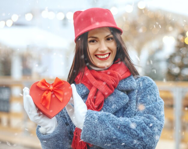 Emotional brunette woman in winter coat holding a gift box at christmas fair during the snowfall