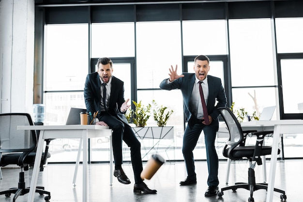 Эмоциональные бизнесмены смотрят на падающий бумажный стаканчик в современном офисе