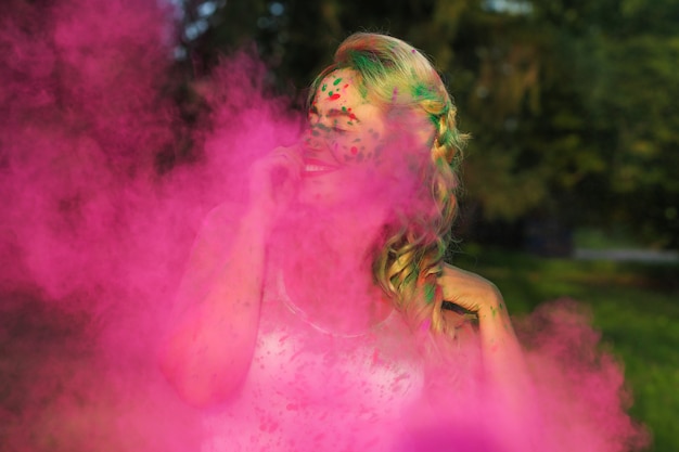 Emotional caucasian woman with curly hair standing n a cloud of pink dry paint, celebrating Holi festival