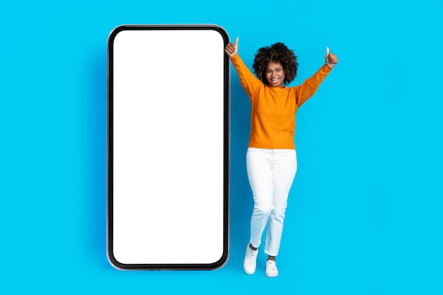 Emotional cheery black lady posing by big smartphone mockup