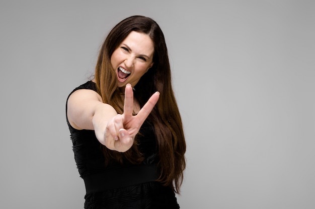 Emotional confident plus size model standing in studio showing peace gesture on gray 
