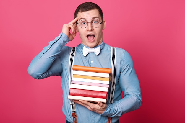 Emotional creative dark haired young man puts forefinger to his temple, holds many books in one hand, opens his mouth and eyes widely, keeping amazing idea in his mind.