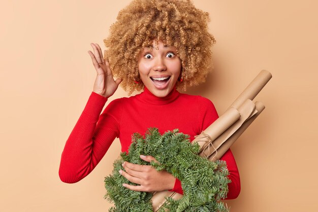 Emotional curly haired cheerful woman keeps hand raised feels happy and surprised exclaims and reacts on awesome news wears red turtleneck going to decorate house before New Year poses indoor