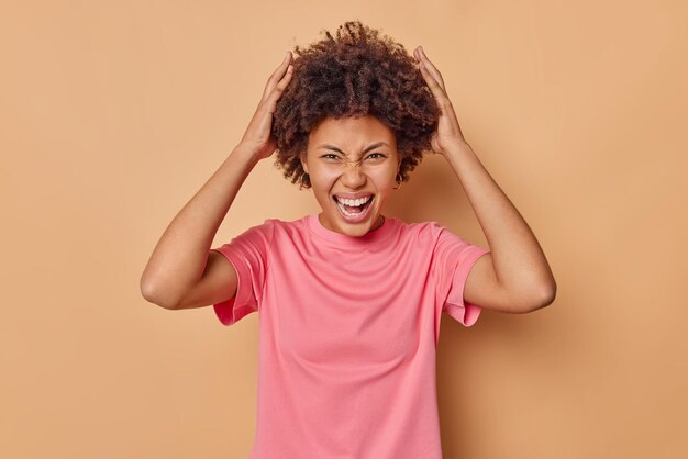 Emotional curly haired woman grabs head exclaims loudly keeps mouth opened reacts on awesome news wears casual pink t shirt isolated over beige backgrounnd realizes her big failure has much work to do