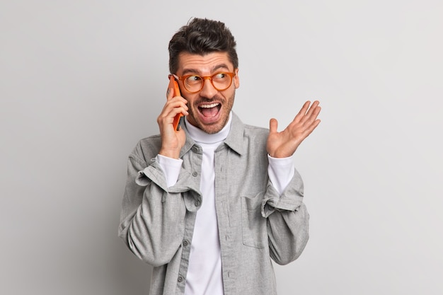 Emotional European man exclaims loudly while talking via mobile phone discusses something actively wears transparent glasses and shirt isolated over grey wall. Technology communication