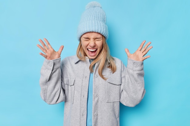 Emotional fair haired young woman exclaims loudly smirks face kees palms raised wears hat and grey jacket reacts on something isolated over blue background People and human reactions concept