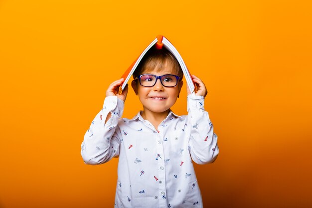 眼鏡をかけて感情的な面白い男の子はカメラに微笑んで、彼の頭、黄色の背景に本を持っています