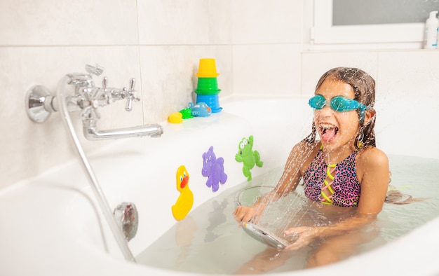 Emotional funny caucasian girl joyfully plays with water while bathing in the bathroom. Concept of entertainment and hygiene of healthy children. Copyspace