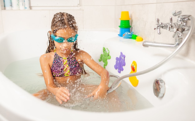 Emotional funny caucasian girl joyfully plays with water while bathing in the bathroom. Concept of entertainment and hygiene of healthy children. Copyspace