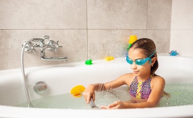 Emotional funny caucasian girl joyfully plays with water while bathing in the bathroom