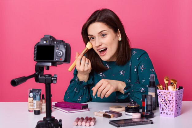 Emotional funny young female sits at table with cosmetics, shooting new make up tutorial for her blog, opening mouth widely, holding professional brush in her hand, wearing flower printed blouse.