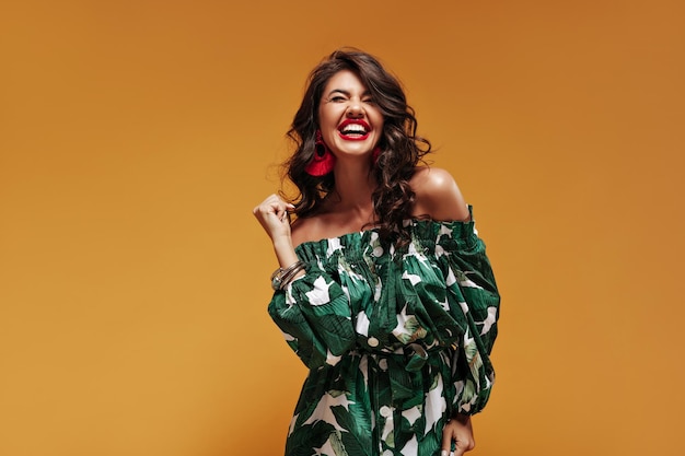 Emotional girl with cute smile and curly wavy hair in green stylish sundress laughing and posing with closed eyes on isolated backdrop