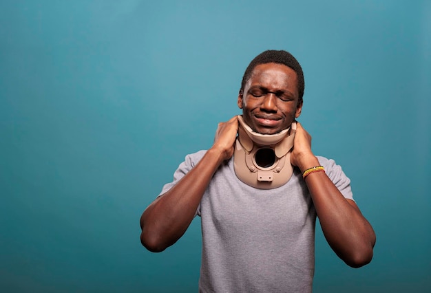 Emotional guy wearing cervical collar to cure physical injury after sprained bone accident. Young man with neck foam suffering from fractured vertebrae, being in pain at rehabilitation.