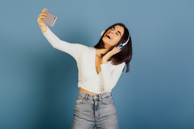 Emotional happy girl with closed eyes wearing white blouse and jeans, standing and making selfie with smile.