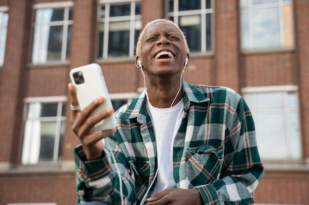 Ragazzo emotivo hipster con acconciatura alla moda utilizzando musica d'ascolto del telefono cellulare, ridendo all'aperto