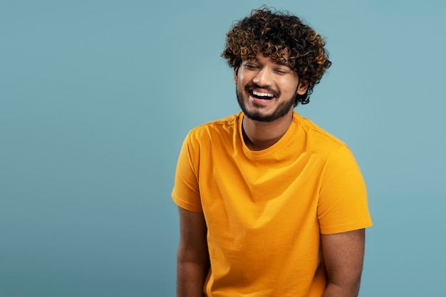 Emotional Indian man with stylish curly hair wearing yellow t shirt laughing with eyes closed