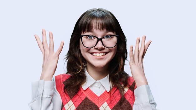 Emotional joyful young female student in glasses with raised hands closeup face on light studio background Emotion joy happiness success surprise