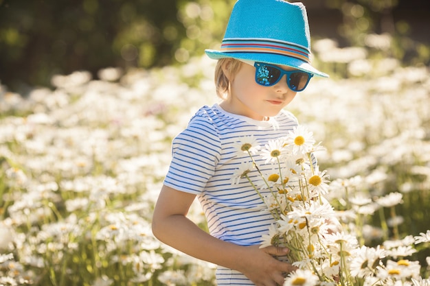 Emotional kid in the camomile field. Cheerful child with flowers wearing hat and sunglasses. Happy boy in the summer time outdoors