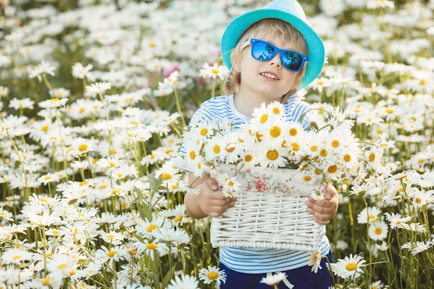 Emotional kid in the camomile field. Cheerful child with flowers wearing hat and sunglasses. Happy boy in the summer time