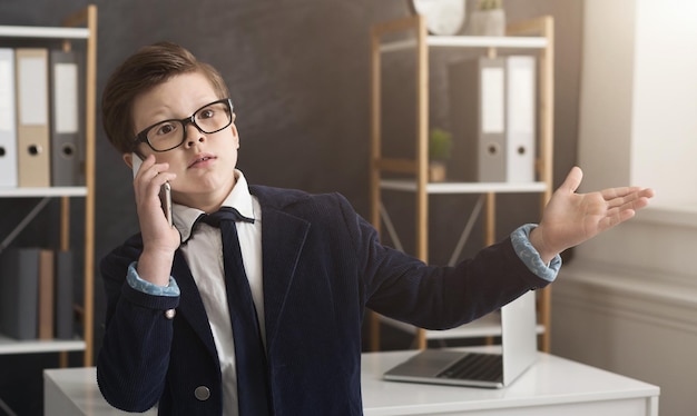 Emotional little boy in suit having serious business talk and gesticulating in office, copy space