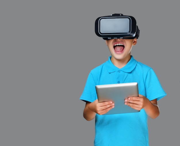 Emotional little boy with vr glasses and tablet PC. Isolated on grey background.