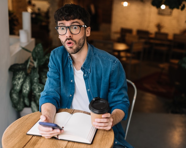 カフェで一杯のコーヒーとスマートフォンを保持している感情的な男