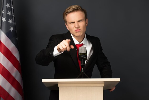 Emotional man pointing with finger on tribune with american flag on black background