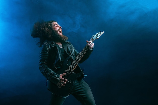 Foto emotivo chitarrista rock uomo con capelli lunghi e barba suona sul colpo di studio di sfondo fumo