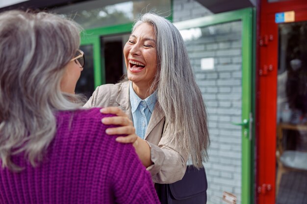 Emotional mature asian lady laughs meeting grey haired best friend on modern city street