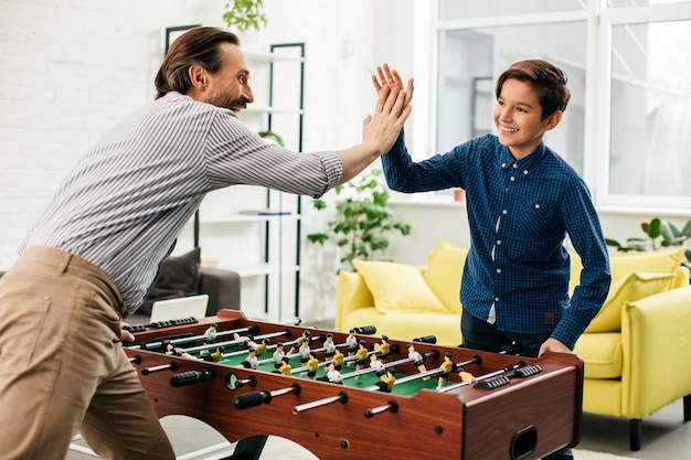 彼の陽気な10代の息子とテーブルサッカーの試合を終え、彼に5を与えながら笑っている感情的な中年の白人男性