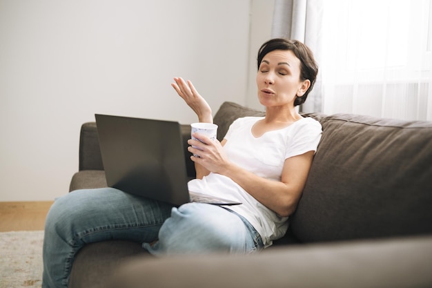 Emotional middle aged woman chatting with online friends Brunette lady with short hair makes video call on sofa