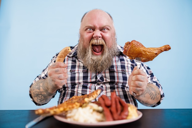 L'uomo obeso emotivo tiene la salsiccia e la coscia di pollo affumicata seduti al tavolo