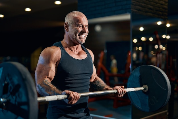 emotional older bald sportsman trains in the gym while pumping up biceps muscles with barbell