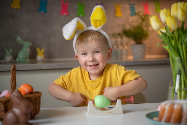 イースターの日にバニーの耳を身に着けている陽気な小さな男の子の感情的な肖像画は、キッチンのテーブルに座ってカラフルなイースターエッグで楽しく笑います