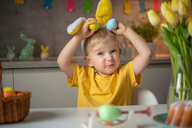 Эмоциональный портрет веселого маленького мальчика с кроличьими ушами в день Пасхи, который смеется, весело играет с красочными пасхальными яйцами, сидя за столом на кухне