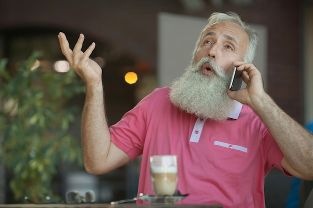 Ritratto emotivo di un uomo allegro ed elegante di affari maturi con la testa calva, con un sorriso che parla sullo smartphone con un amico. uomo anziano barbuto