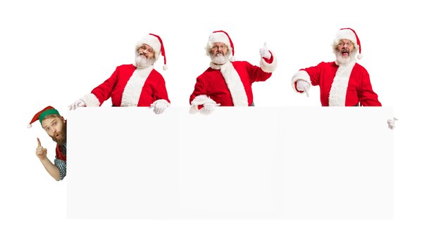 Эмоциональное поздравление Деда Мороза с Новым 2020 годом и Рождеством. Мужчины в традиционных костюмах держат большой белый лист для вашей рекламы. Зима, праздничное настроение, подарки, пожелания. Copyspace.