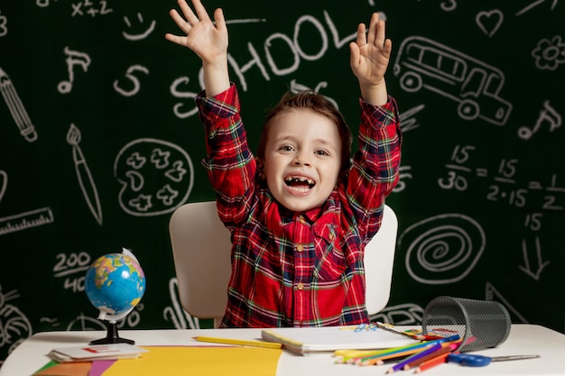 多くの学用品を机の上に座っている感情的な学校の男の子。学校の最初の日。小学校の子供男の子。学校に戻る。小学校の子。