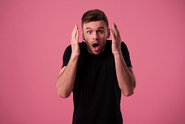 Emotional and shocked face of young man in black t-shirt isolated on pink wall