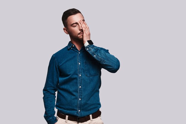 Emotional stress. Frustrated young man touching his face with hand while standing against grey background