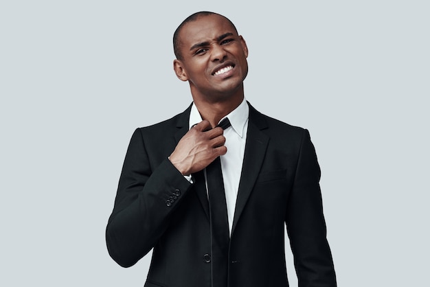 Emotional stress. Tired young African man in formalwear taking off necktie while standing against grey background