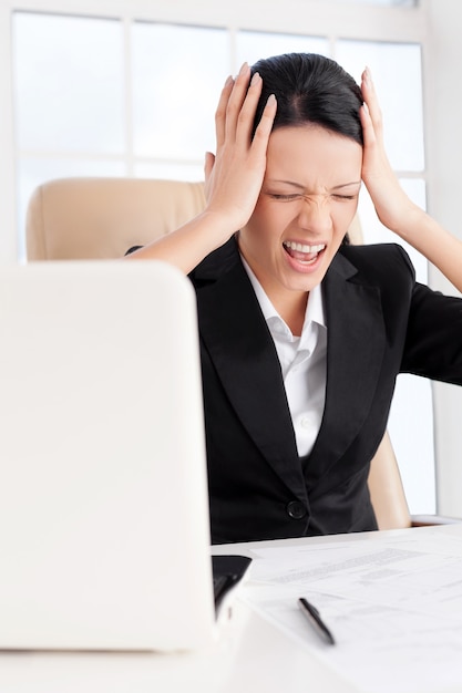 Emotional stress. Young business woman holding head in hands and shouting while sitting at her working place