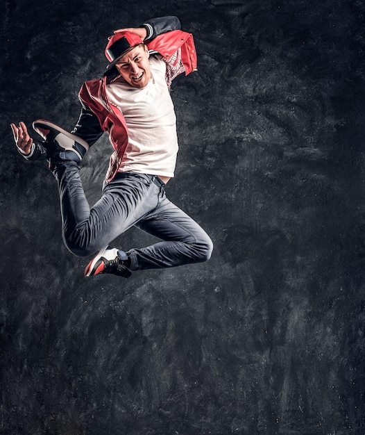 Emotional stylish dressed guy performing break dance jumping. Studio photo against a dark textured wall