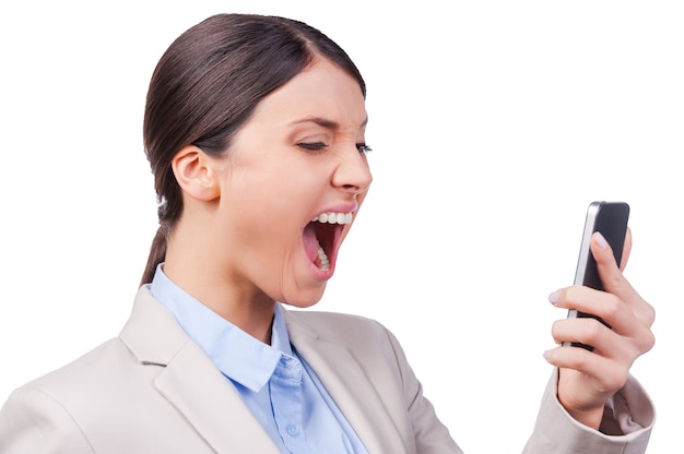 Emotional talking. Beautiful young businesswoman holding mobile phone and screaming while standing against white background