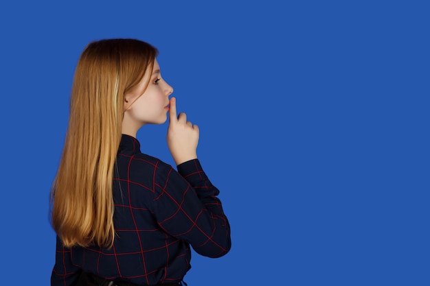 Emotional thirteen-year-old cute teenage girl in glasses with finger at mouth looks carefully away, isolated against blue background. Caucasian child put finger to lips in sign of silence. Copy space