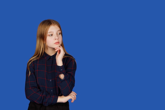 Emotional thirteen-year-old cute teenage girl thinking and looking away, isolated against blue Royal background. Caucasian child in black clothes shows question mark emotions. Copy space