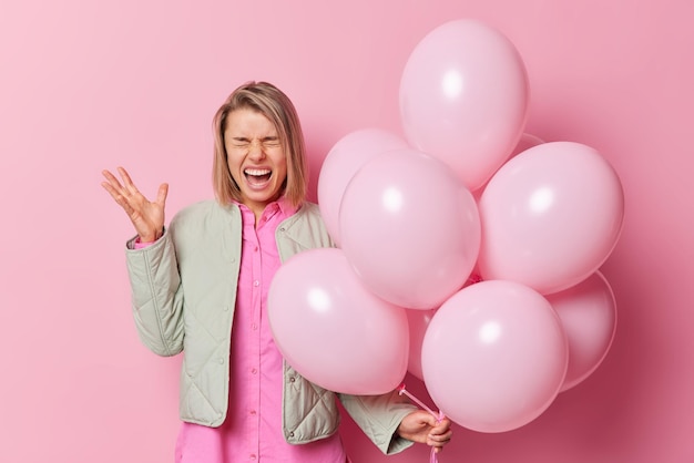 Emotional woman screams outraged shouts at someone holds bunch of inflated balloons keeps hand raised wears shirt and jacket expresses negative emotions angry to hear annoying music on party