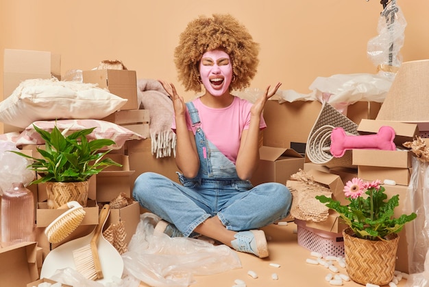 Emotional woman with curly hair applies beauty mask raises arms\
and exclaims loudly wears t shirt and denim overalls sits crossed\
legs on floor in new apartment packs personal things in boxes