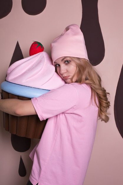 분홍색 티셔츠와 모자를 쓰고 큰 컵케이크 달콤한 장식으로 포즈를 취한 감정적인 젊은 금발 모델